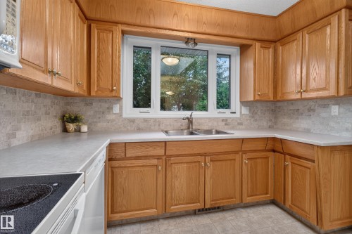 Kitchen featuring extensive wood cabinetry, light-toned countertops, a stainless steel double-basin sink with a gooseneck faucet, a white built-in dishwasher, and a grey tile backsplash - 17603 85 Avenue Nw, Edmonton, AB 
