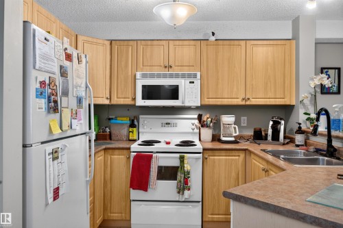 Functional kitchen featuring light wood-finish cabinetry and a white appliance suite including a built-in microwave and refrigerator - 414 11214 80 Street, Edmonton, AB 