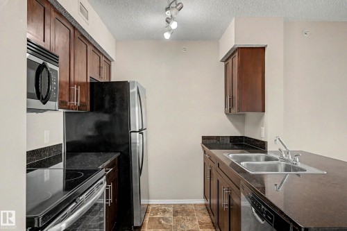 The kitchen features dark wood cabinetry, black countertops, stainless steel appliances including a microwave and refrigerator, and a double basin stainless steel sink - 404 1080 Mcconachie Boulevard, Edmonton, AB 