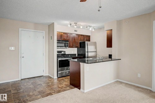 Kitchen area featuring dark wood-finish cabinetry, stainless steel appliances, and a black countertop with an integrated sink and breakfast bar - 404 1080 Mcconachie Boulevard, Edmonton, AB 