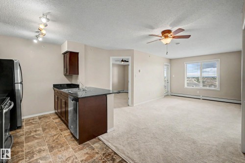 Open-concept living space featuring a kitchen with dark cabinetry, stainless steel appliances, a black countertop, and tile flooring - 404 1080 Mcconachie Boulevard, Edmonton, AB 
