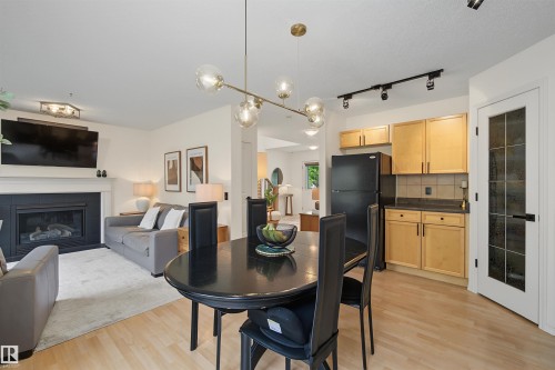 Open-concept living area featuring light wood-finish flooring, a contemporary fireplace with a white mantel, and a kitchen area with light wood cabinetry - 15 450 Hyndman Crescent, Edmonton, AB 