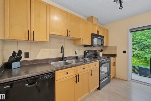 Kitchen featuring light wood cabinetry with dark hardware, black appliances, a tile backsplash, and dark countertops - 15 450 Hyndman Crescent, Edmonton, AB 