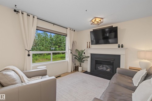 Living area featuring a wide window with exterior tree scenery, a built-in fireplace with a black surround and white mantel, and light-toned walls - 15 450 Hyndman Crescent, Edmonton, AB 