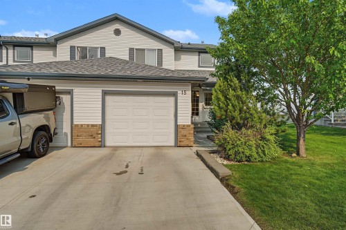 Light-colored siding exterior featuring a multi-pane garage door, a concrete driveway, a front entry with visible steps, and established landscaping including a mature tree and shrubs - 15 450 Hyndman Crescent, Edmonton, AB 