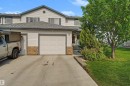 Light-colored siding exterior featuring a multi-pane garage door, a concrete driveway, a front entry with visible steps, and established landscaping including a mature tree and shrubs - 15 450 Hyndman Crescent, Edmonton, AB 