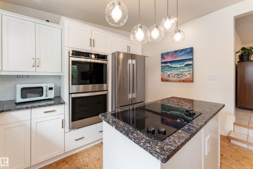 Kitchen featuring white cabinetry, stainless steel double wall ovens, a side-by-side refrigerator, a cooktop integrated into a kitchen island, and multiple pendant lights - 7816 142 Street, Edmonton, AB 