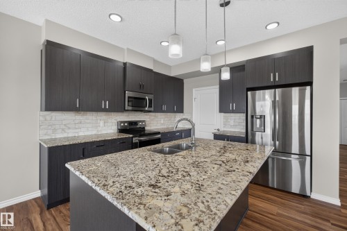 Contemporary kitchen featuring a large island with granite countertop and dual basin sink, dark wood-finish cabinetry, stainless steel appliances, and a tiled backsplash - 2460 15 Avenue, Edmonton, AB 