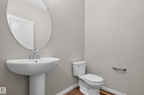 Bathroom featuring a white pedestal sink with chrome fixtures, an oval mirror, a white toilet, and wood-finish flooring - 2460 15 Avenue, Edmonton, AB 