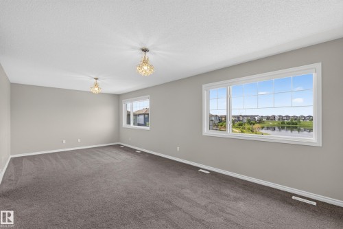 Expansive room featuring a large window with a water backdrop, neutral gray wall paint, textured gray carpeting, and two ornate crystal ceiling fixtures - 2460 15 Avenue, Edmonton, AB 