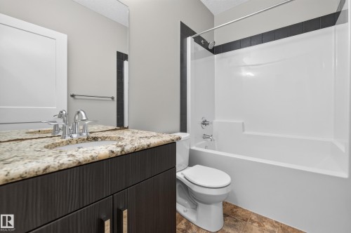 Bathroom featuring a dark wood-finish vanity with a light speckled countertop, an undermount sink with chrome fixtures, a shower-tub combination with a white surround and black tile accent, and a light-colored tile floor - 2460 15 Avenue, Edmonton, AB 