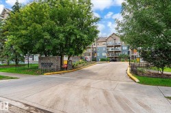 Scona Gardens entrance featuring a stone monument with wrought iron fencing, surrounded by mature trees and manicured landscaping, with a multi-story residential building in the background - 