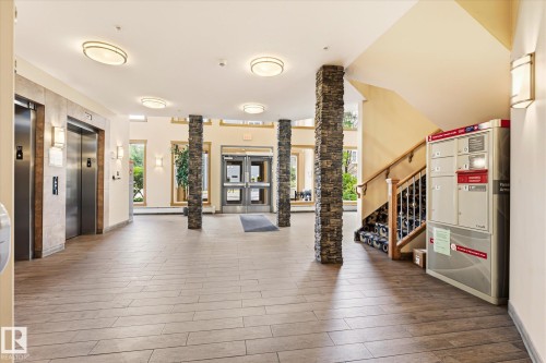 Spacious building lobby featuring wood-finish tile flooring, stacked stone columns, multiple elevators, and a visible staircase with a wooden handrail - 402 10121 80 Avenue, Edmonton, AB 