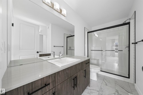 Bathroom featuring a spacious vanity with wood-finish cabinetry, a large integrated mirror, a quartz-finish countertop, and a walk-in shower with a black-framed glass enclosure - 2953 Coughlan Green Green, Edmonton, AB 
