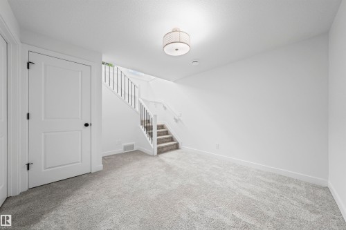 Spacious room featuring light gray carpet, white walls, a flush-mount ceiling light, a two-panel door with black hardware, and a staircase with a black metal spindle railing - 2953 Coughlan Green Green, Edmonton, AB 