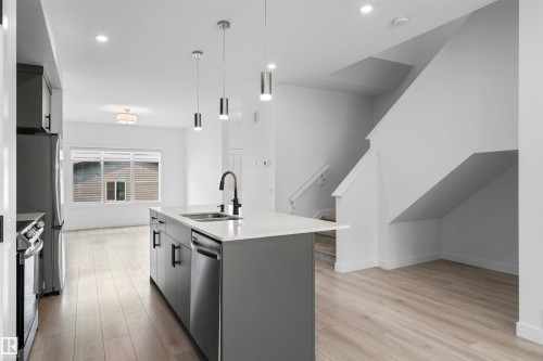 Bright open-concept kitchen featuring a central island with a sink and pendant lighting, stainless steel appliances, modern cabinetry, and wood-finish flooring - 2953 Coughlan Green Green, Edmonton, AB 