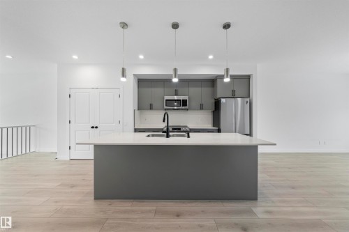 Modern kitchen featuring a large central island with a solid surface countertop, matte gray cabinetry, stainless steel appliances, and wood-finish flooring - 2953 Coughlan Green Green, Edmonton, AB 