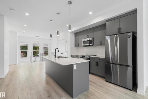Contemporary kitchen featuring an island with a sink, stainless steel appliances, dark cabinetry, light countertops, and light wood-finish flooring - 2953 Coughlan Green Green, Edmonton, AB 