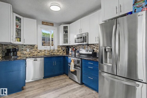 Kitchen featuring wood-finish flooring, two-tone cabinetry with white upper and blue lower cabinets, granite-style countertops, and a mosaic tile backsplash - 3622 115 Avenue, Edmonton, AB 