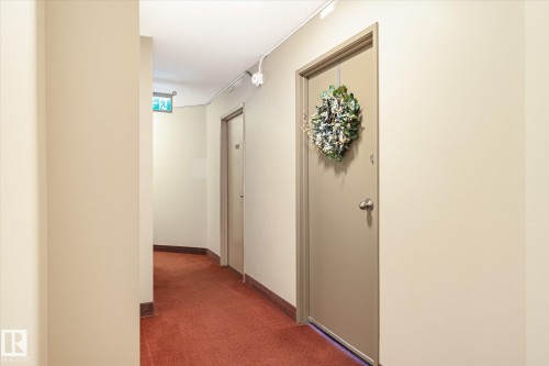Neutral-toned walls complement the red carpet flooring in this hallway, featuring a robust entry door with a simple doorknob - 503 10045 117 Street, Edmonton, AB 