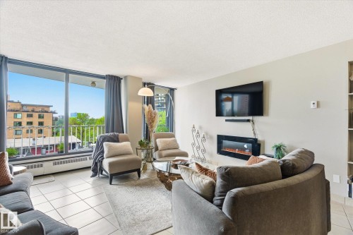 Living area featuring a full-width window wall, white square tile flooring, a wall-mounted electric fireplace, and a built-in shelving unit - 503 10045 117 Street, Edmonton, AB 