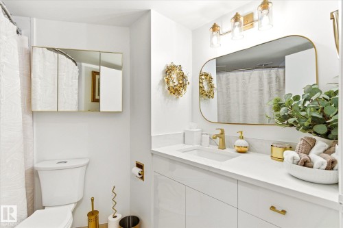 Bathroom featuring a single vanity with a white countertop and integrated sink, gold-toned faucet, and a rectangular mirror with a gold-toned frame - 503 10045 117 Street, Edmonton, AB 