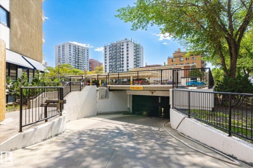 Building exterior featuring a concrete entrance ramp leading to an underground parking garage, surrounded by black metal railings and mature trees - 503 10045 117 Street, Edmonton, AB 