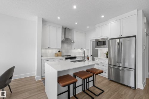 Contemporary kitchen featuring white cabinetry, a waterfall island with a sink, stainless steel appliances, and wood-finish flooring - 20907 131 Avenue, Edmonton, AB 