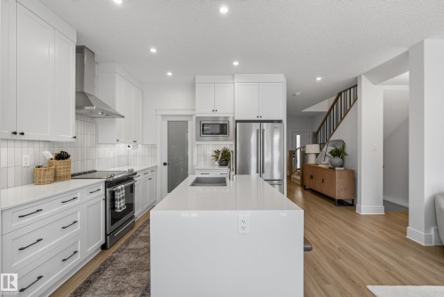 Modern kitchen featuring white cabinetry, stainless steel appliances, a white tile backsplash, and an expansive center island with an undermount sink - 20907 131 Avenue, Edmonton, AB 