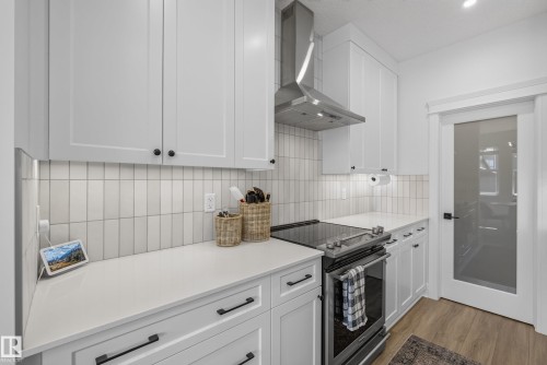Modern kitchen featuring white shaker cabinetry, stainless steel range hood, sleek electric range, light-toned countertops, and vertical tile backsplash - 20907 131 Avenue, Edmonton, AB 