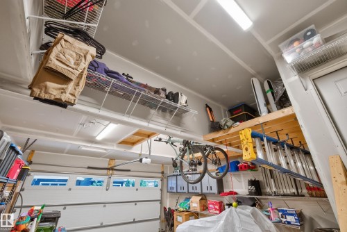 Spacious garage featuring a paneled garage door with upper window inserts, overhead fluorescent lighting, high ceilings, built-in wire shelving, and a robust wooden storage platform - 20907 131 Avenue, Edmonton, AB 
