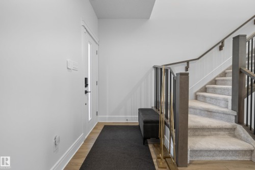 Entryway featuring a partially glass-paneled door, wood-finish flooring, and a carpeted staircase with dark wood-finish newel posts and handrails, complemented by black and gold-tone balusters - 20907 131 Avenue, Edmonton, AB 
