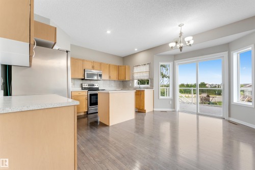 Open-concept kitchen and dining area featuring wood-finish flooring, a central kitchen island with a light-toned countertop, stainless steel appliances, and ample natural light from multiple windows and a sliding glass door - 3449 28 Street, Edmonton, AB 