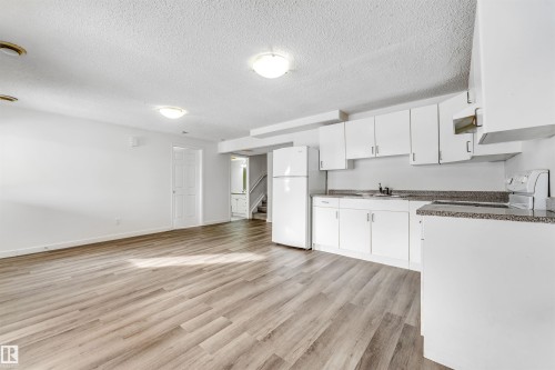 Open-concept living space with wood-finish flooring, a full kitchen featuring white cabinetry, a refrigerator, and speckled countertops - 3449 28 Street, Edmonton, AB 