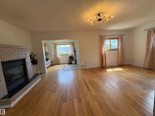 Living Room, wood-finish flooring, a brick fireplace with a dark stone hearth, and multiple windows with a view of the front entrance. - 4806 50 Avenue, Elk Point, AB 