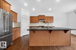 Kitchen featuring a large island with a dark countertop, wood-finish cabinetry, stainless steel appliances, tile backsplash, and wood-finish flooring - 