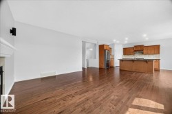 Open-concept living space featuring wood-finish flooring, a kitchen with wood cabinetry and stainless steel appliances, a kitchen island with a breakfast bar, recessed lighting, and a fireplace with a white mantel and dark hearth - 