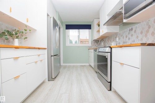 Contemporary kitchen featuring white cabinetry with gold-tone hardware, wood-finish countertops, stainless steel appliances, a patterned tile backsplash, and light wood-finish flooring - 3531 46 Street, Edmonton, AB 