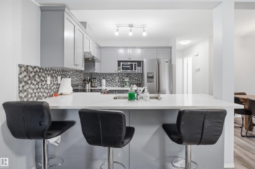 Modern kitchen featuring a breakfast bar with a white countertop, gray flat-panel cabinetry, a mosaic tile backsplash, and stainless steel appliances - 92 230 Edwards Drive, Edmonton, AB 