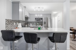 Modern kitchen featuring a breakfast bar with a white countertop, gray flat-panel cabinetry, a mosaic tile backsplash, and stainless steel appliances - 