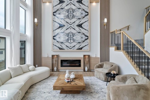 Double-height living space featuring expansive windows, a built-in electric fireplace, wood-finish wall paneling, and an open staircase with metal balusters - 13152 187 Avenue, Edmonton, AB 