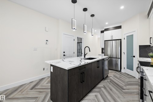 Modern kitchen featuring an island with a stone-look countertop and integrated sink, herringbone wood-finish flooring, stainless steel appliances, contemporary pendant lighting, and a frosted glass door - 8319 81 Avenue Nw, Edmonton, AB 