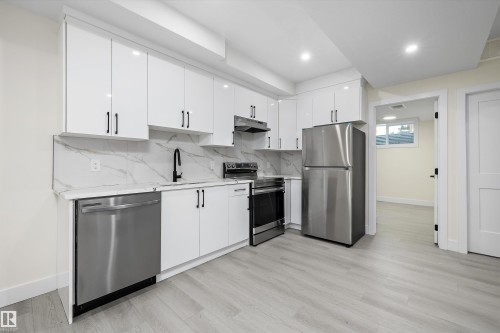 Modern kitchen featuring white cabinetry with black hardware, a stainless steel appliance suite, light-toned wood-finish flooring, and a stone-patterned backsplash - 8319 81 Avenue Nw, Edmonton, AB 