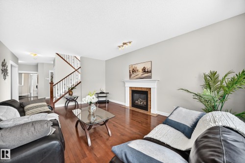 Spacious living area featuring rich wood-finish flooring, a contemporary fireplace with a white mantel and tile surround, and a visible staircase with dark wood railings - 1330 114B Street, Edmonton, AB 
