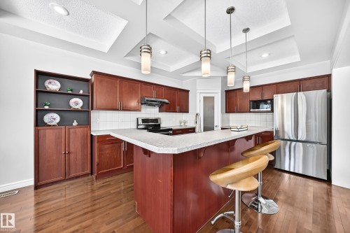 Kitchen featuring wood-finish flooring, a tiered tray ceiling with recessed lighting and pendant fixtures, cherry wood cabinetry, stainless steel appliances, and a central island with a light-colored countertop - 1330 114B Street, Edmonton, AB 