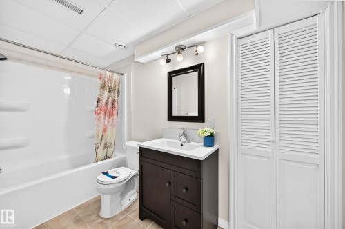 Bathroom featuring a white tub/shower combination, a dark wood-finish vanity with a white countertop and integrated sink, a dark-framed mirror, and bi-fold louvered doors - 1330 114B Street, Edmonton, AB 