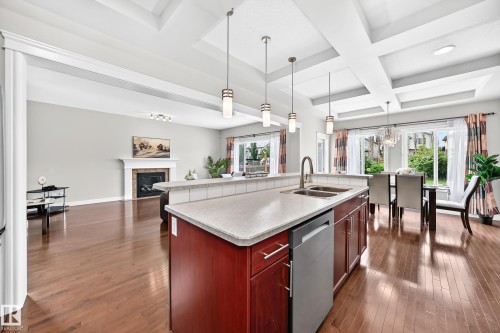 Open-concept living area featuring rich wood-finish flooring, a contemporary kitchen island with a double sink and integrated dishwasher, coffered ceilings, a fireplace with a mantel, and large windows - 1330 114B Street, Edmonton, AB 