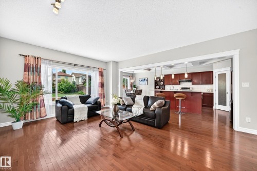 Open-concept living area featuring rich wood-finish flooring, a large picture window, and a kitchen with warm-toned cabinetry, a breakfast bar, and pendant lighting - 1330 114B Street, Edmonton, AB 