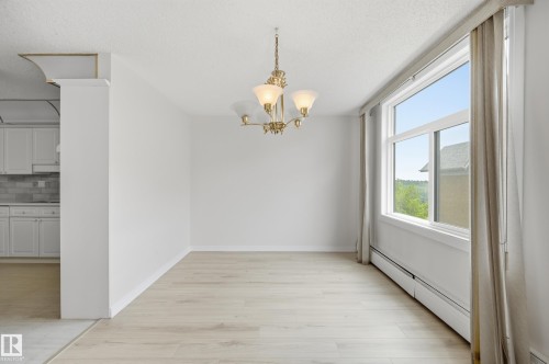 Spacious room featuring wood-finish flooring, a large window, a brass chandelier, and white baseboards - 504 11710 100 Avenue, Edmonton, AB 