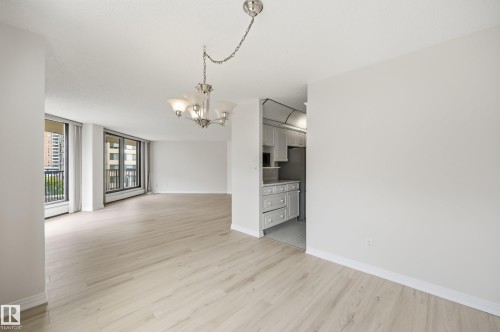 Open-concept living area featuring light wood-finish flooring, ample natural light from large windows, a brushed nickel chandelier, and recessed kitchen entry - 504 11710 100 Avenue, Edmonton, AB 